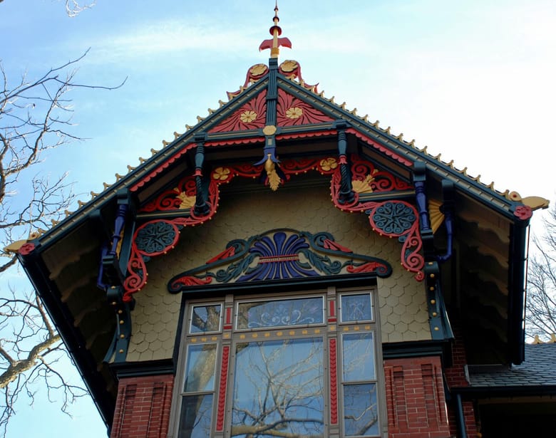 Top of a brown building with shingled siding and colorful wood carvings around a window.