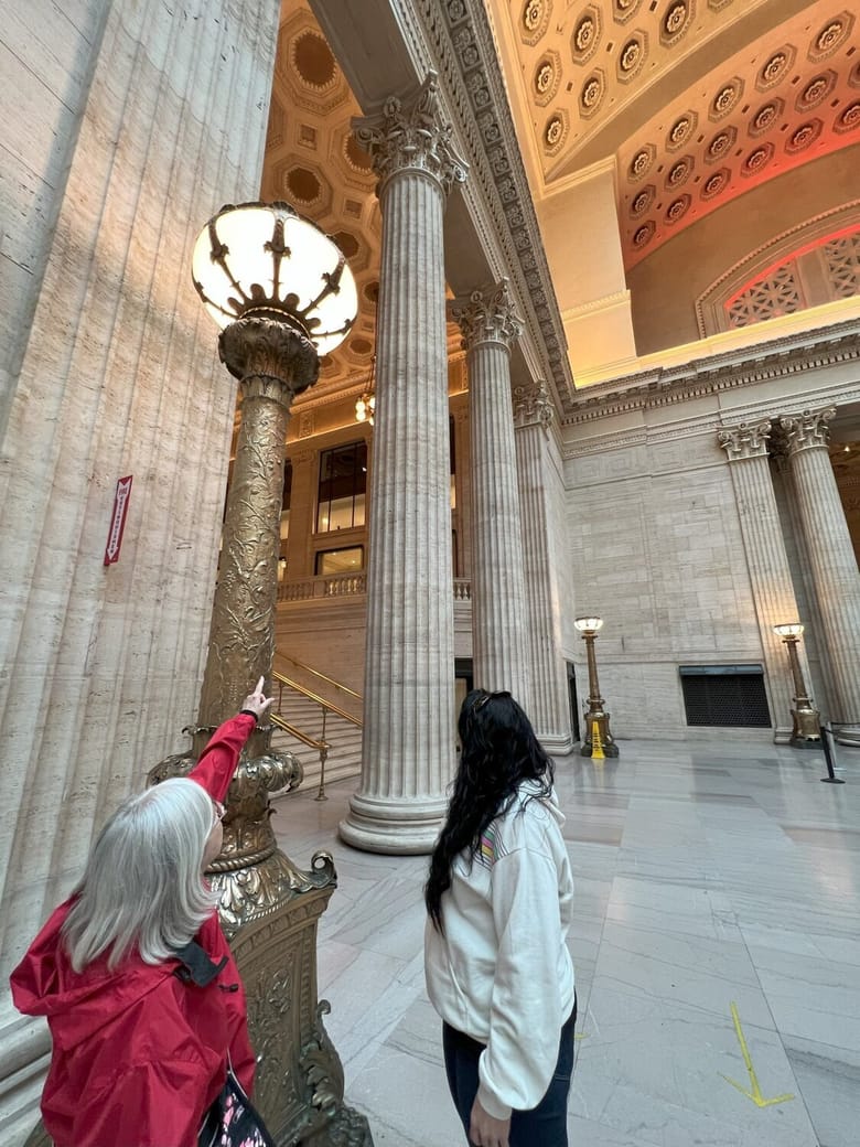 Photo of a docent and tour goer. The docwent is pointing at a golden light post and there are columns and a valuted ceiling in the background.