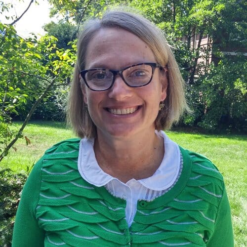 Headshot of a smiling caucasian woman with green sweater and framed glasses