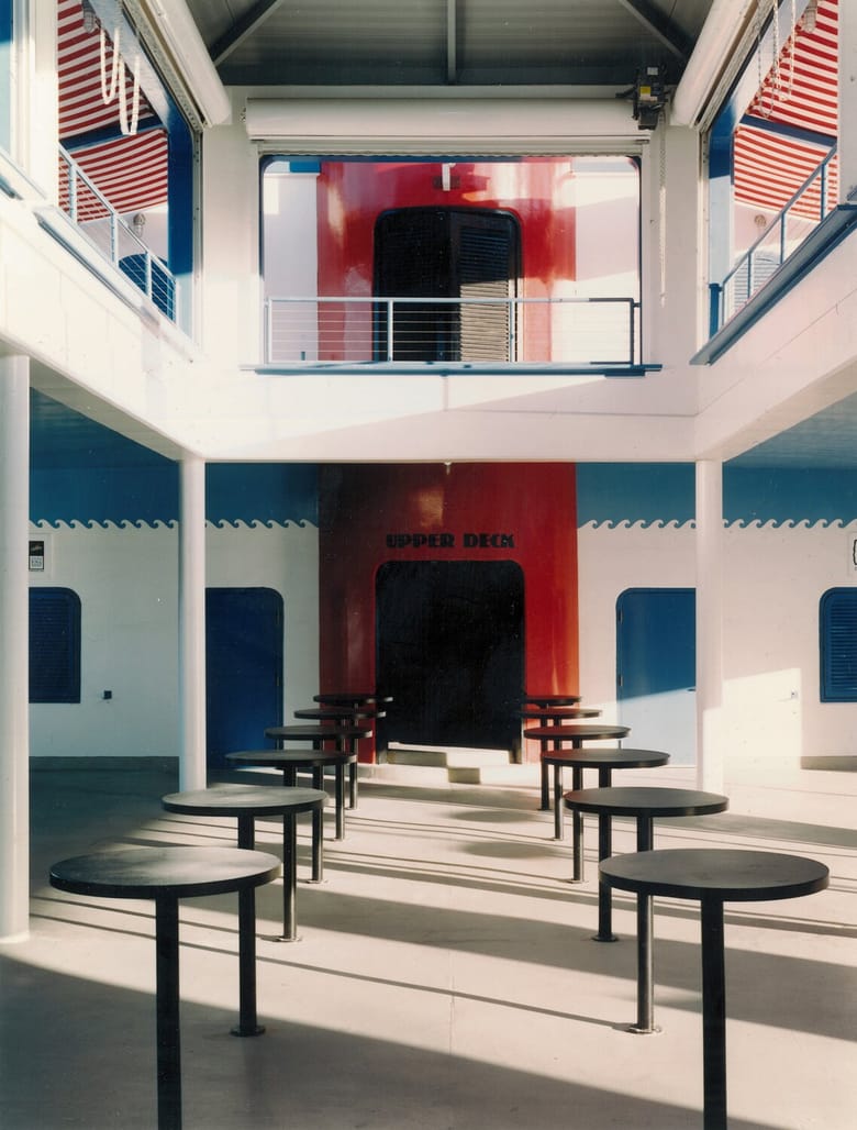 An interior view of the North Avenue Beach boathouse, showcasing its multi-level, ship-inspired architecture. The ground floor features numerous round, dark tables arranged in rows. White pillars support the upper levels. In the center, a large, dark opening is labeled "UPPER DECK" in red text, leading to stairs or a ramp. The walls are white with blue trim and a decorative blue wave pattern. On the upper level, a red wall with a large black opening is visible, resembling the "smokestack" exterior. Red and white striped awnings or coverings are visible on either side of the upper level, extending towards the viewer.