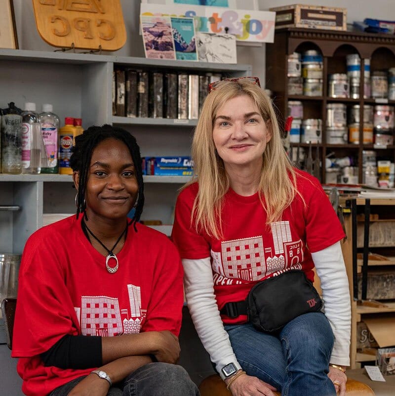 Two ladies in red volunteer short seated and smiling at the camera