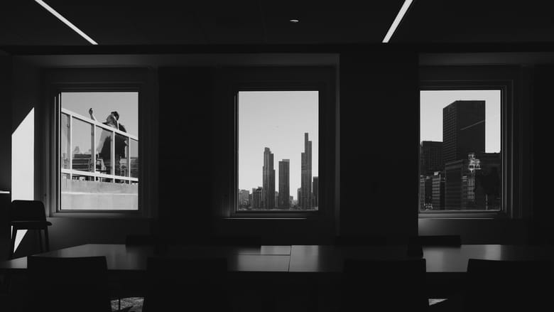 Black and white image in a dark room with three bright tall rectangular windows the two on the far right feature tall buildings and the one on the left features a couple taking a picture together.