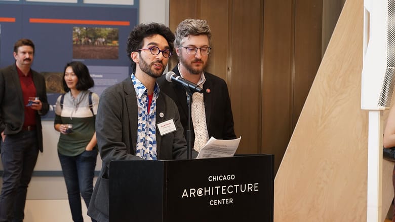 Two men speaking at a podium inside a gallery space at an event