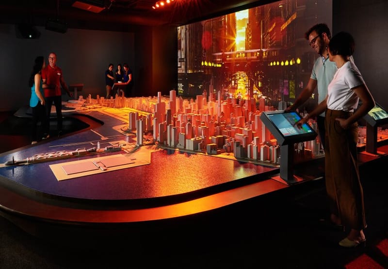 Two people in a red lit room looking at a model of the city