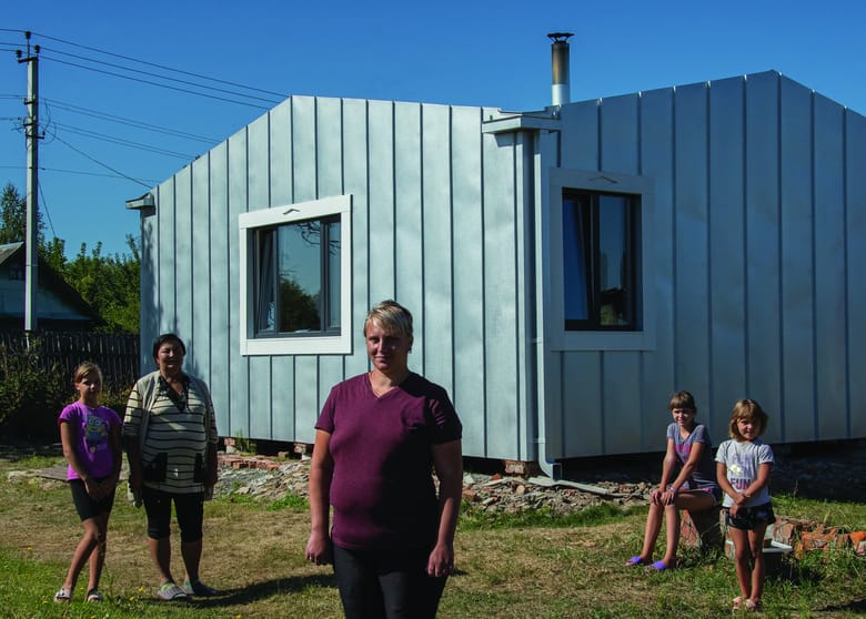 Image of a woman with short blone hair standing in the foreground with two children to her right and a woman and child to her left. They all stand in front of a metal one story building.