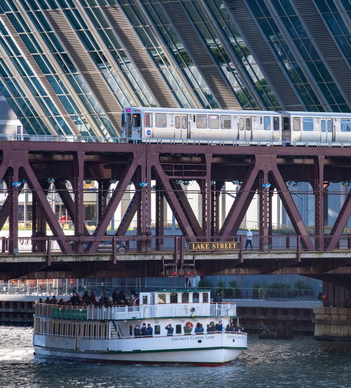 A river cruise going under a bridge with a train going over the bridge