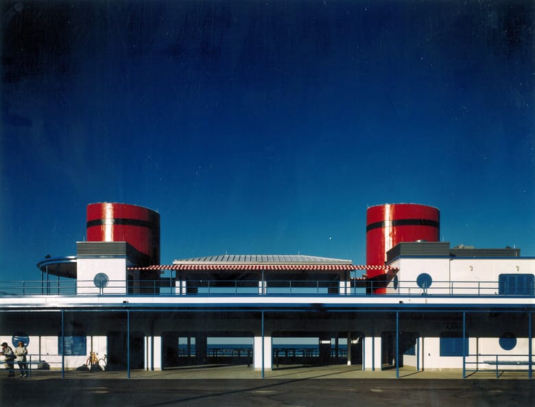 A vibrant, low-angle shot of the North Avenue Beach boathouse in Chicago under a deep blue sky. The building, designed to resemble a ship, features white walls, blue railings, and two prominent, red-banded cylindrical structures on its roof, mimicking smokestacks. A striped awning covers a central outdoor area between the two "smokestacks." The lower level of the building is open, revealing glimpses of Lake Michigan through its arched supports. Two people and a bicycle are visible on the left side of the lower level. The intense blue of the sky contrasts sharply with the white and red of the building.