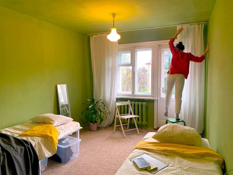Woman stands on a green stool in a room with lime green walls adjusting white curtains.