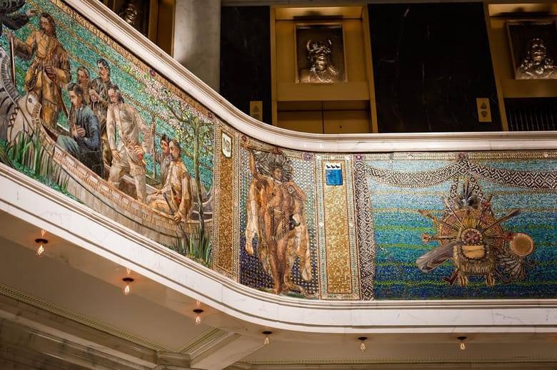 Mosaic on the side of a balcony indoors depicting a panel of men standing in a boat and a panel of a single man standing and wearing a head dress.