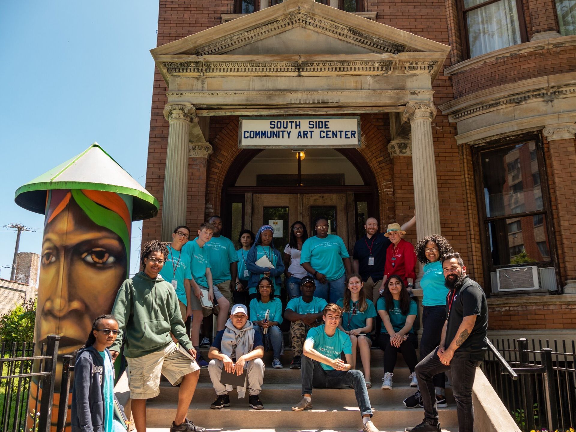 Group of teens sitting on the stairs in front of a red brick building all wearing teal shirts.