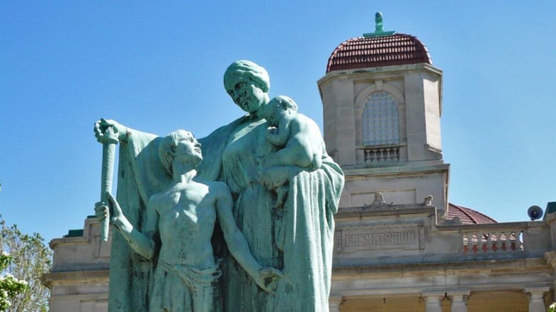 A green sculpture of a woman holding a toddler and a child in front of a large grey building in the background amidst a cemetary