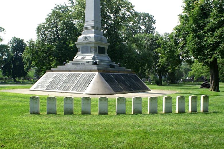 Large white monument with 12 smaller grave markers on grassy plain.