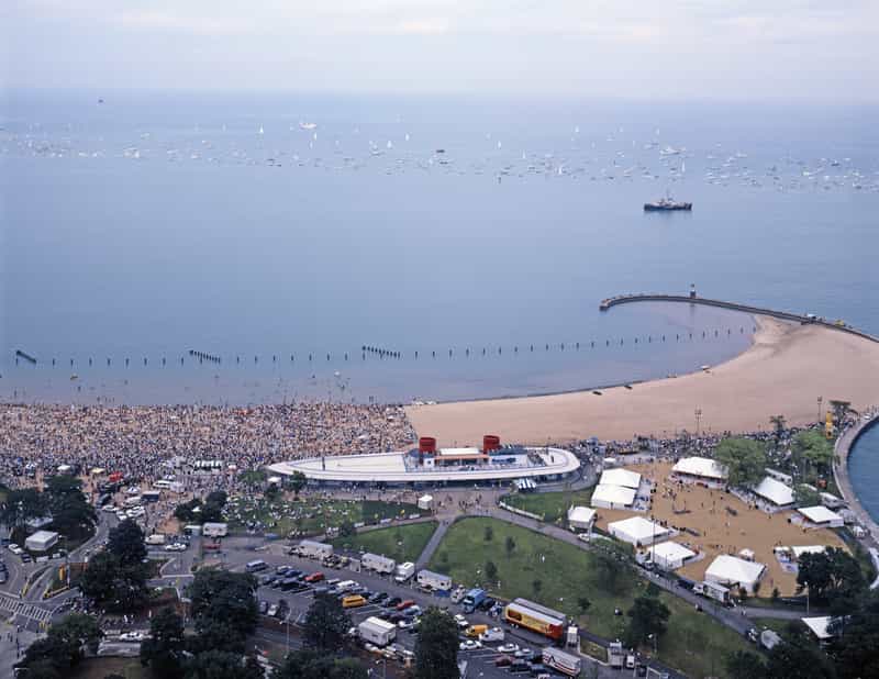 An aerial shot shows a crowded beach along a large body of water, likely Lake Michigan, with numerous sailboats dotting the horizon under a pale sky. In the foreground, a sandy beach is packed with people. Adjacent to the beach is a modern, boat-shaped building with red accents on its roof. To the right of the building, a large area with multiple white tents is set up on what appears to be muddy ground, possibly indicating recent rain or a specific event setup. A paved road with parked cars and a few buses curves around a grassy area in the lower left. A long pier extends into the water from the right, ending at a small lighthouse. In the distance, a large ship is visible on the water.