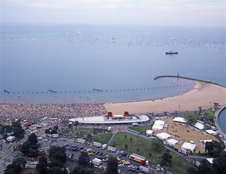 An aerial shot shows a crowded beach along a large body of water, likely Lake Michigan, with numerous sailboats dotting the horizon under a pale sky. In the foreground, a sandy beach is packed with people. Adjacent to the beach is a modern, boat-shaped building with red accents on its roof. To the right of the building, a large area with multiple white tents is set up on what appears to be muddy ground, possibly indicating recent rain or a specific event setup. A paved road with parked cars and a few buses curves around a grassy area in the lower left. A long pier extends into the water from the right, ending at a small lighthouse. In the distance, a large ship is visible on the water.