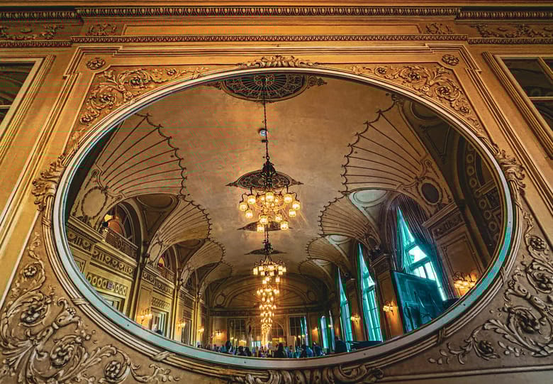 Image of a mirror reflecting a long row of light fixtures and an ornate ceiling in a large building.