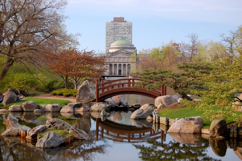 A serene Japanese garden with a winding stone pathway bordered by lush greenery, leading to a tranquil pond reflecting overhanging trees and a pagoda-style structure in the background.