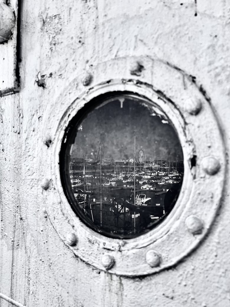 Black and white image looking into a rusted and weathered boat porthole that reflects the ferris wheel of Navy Pier.