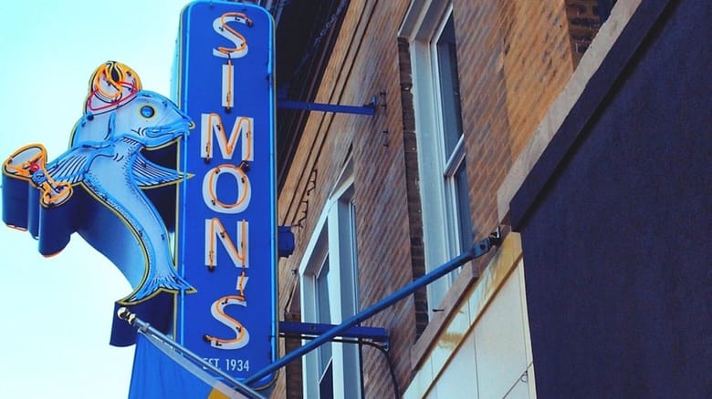 Vertical sign hanging from brick building. Blue background with white letters spelling Simon's.