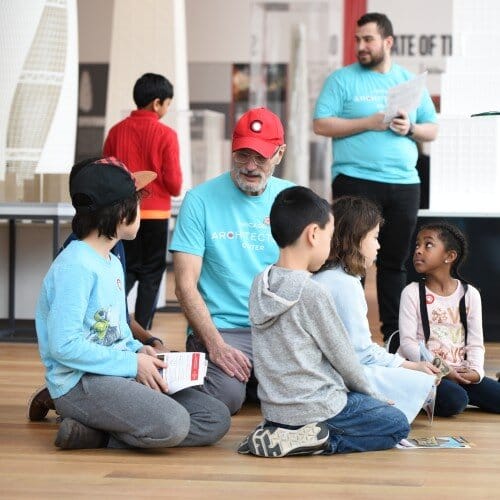 Children kneeling on the floor with workbooks talking with a person in a teal t-shirt and red hat. They have a short white beard.