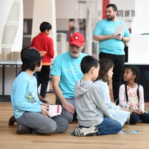 Children kneeling on the floor with workbooks talking with a person in a teal t-shirt and red hat. They have a short white beard.