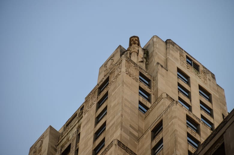 Exterior view of angled beige building with many windows and a statue on the top corner