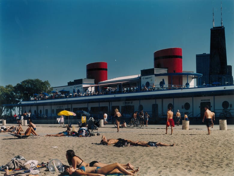 A sunny, mid-day shot of North Avenue Beach in Chicago, featuring the distinctive boathouse building in the background. The sandy beach in the foreground is populated with sunbathers lying on towels and chairs. A yellow beach umbrella stands out in the middle ground. The boathouse, designed like a ship, has white walls, blue trim, and two prominent red cylindrical structures on its roof. People are visible on the boathouse's upper deck and on the ground level. In the far background, the top of the John Hancock Center (now 875 North Michigan Avenue) is visible, along with other city buildings, under a clear blue sky.