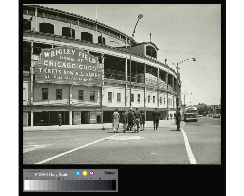 Black and white image of the exterior of Wrigley Field with a large sign that reads "Wrigley Field Home of the Chicago Cubs" as the focal point, and people walking towards the stadium in the foreground.